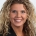 A woman with blond curly hair is wearing a dark jacket and smiling at the camera