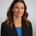 A woman with curly long brown hair is wearing work attire and posing for the camera