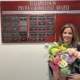 A woman holding a flower bouquet is posing in front of a plaque board.