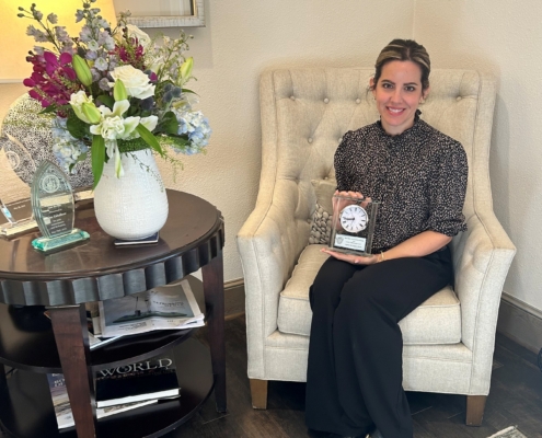 A woman wearing business attire is sitting in a white chair holding an award.