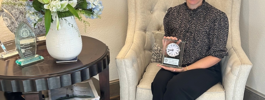 A woman wearing business attire is sitting in a white chair holding an award.