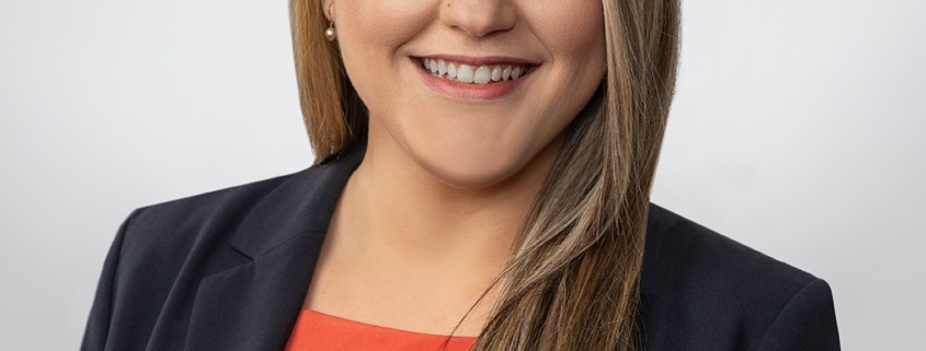 A woman with long brown hair is wearing a red top and a black jacket and smiling at the camera